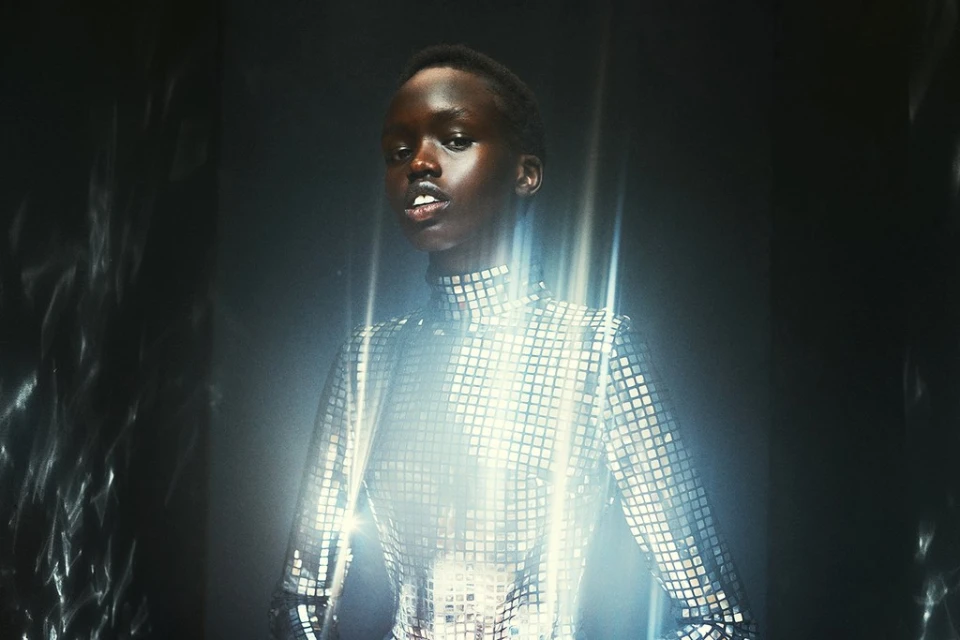 Stylish woman in a shimmering, square-patterned turtleneck dress poses indoors with a faint smile, creating a striking, modern aesthetic.