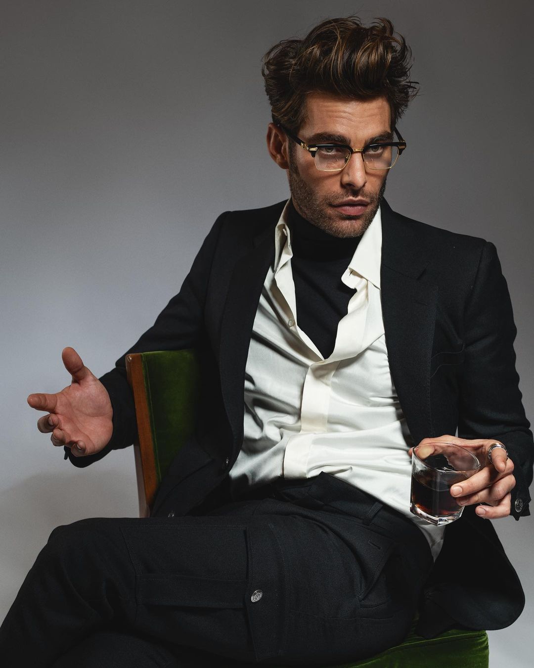 Man sitting in chair wearing glasses, black blazer and pants, holding a glass of alcohol in a studio with grey background.