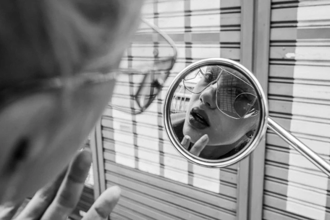 Black and white close-up of a woman reflected in a motorcycle mirror applying lip gloss, with a striped garage door in the background.