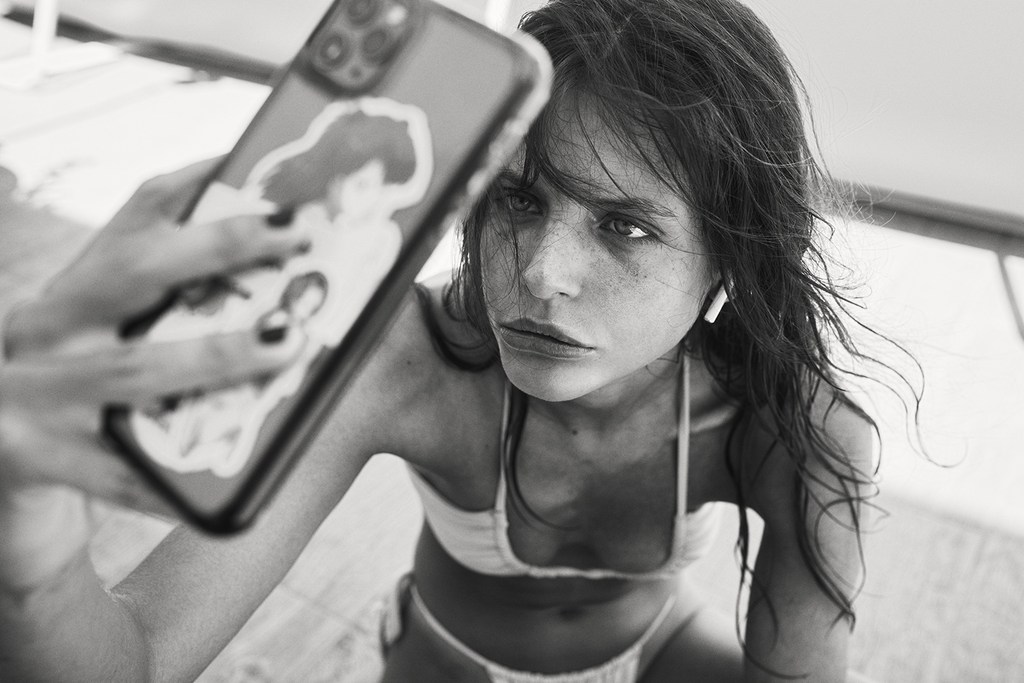 Monochrome selfie of a young adult woman in a bikini, holding a phone with stickers. The image has a shallow depth of field.