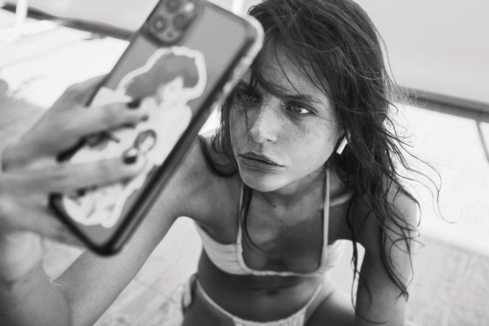 Monochrome selfie of a young adult woman in a bikini, holding a phone with stickers. The image has a shallow depth of field.