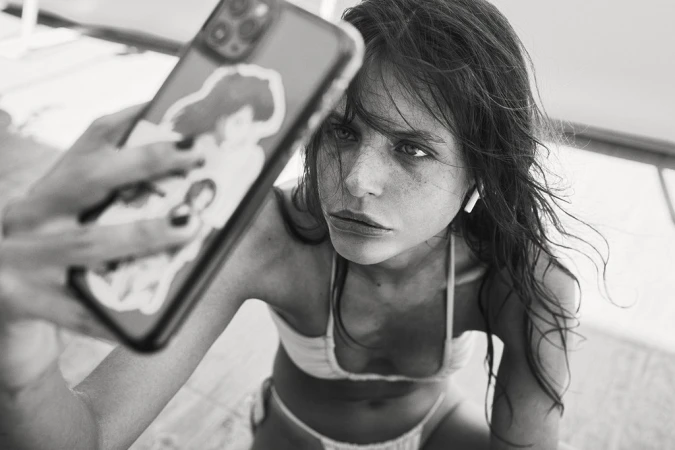 Monochrome selfie of a young adult woman in a bikini, holding a phone with stickers. The image has a shallow depth of field.