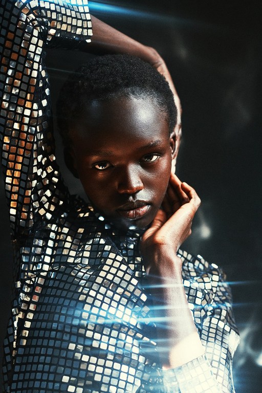 Headshot of an African American woman with hands near her face, wearing a reflective tile-patterned shirt against a dark background.