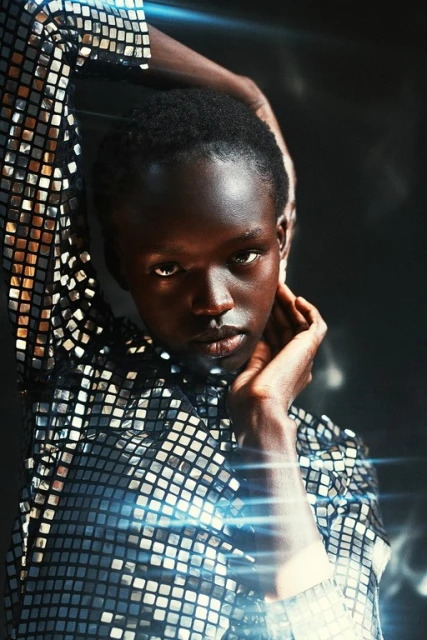 Headshot of an African American woman with hands near her face, wearing a reflective tile-patterned shirt against a dark background.