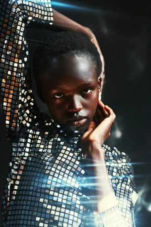 Headshot of an African American woman with hands near her face, wearing a reflective tile-patterned shirt against a dark background.