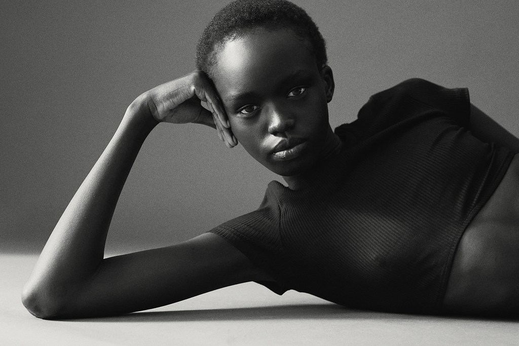 Portrait of a black woman with short hair wearing a dark short-sleeve top lying down and resting her head on her hand against a grey backdrop.