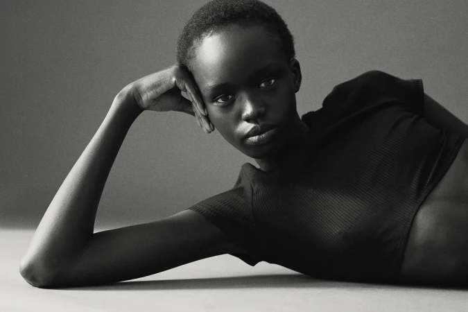 Portrait of a black woman with short hair wearing a dark short-sleeve top lying down and resting her head on her hand against a grey backdrop.