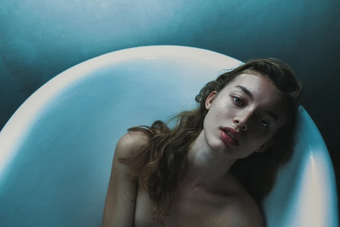 Close-up of a young woman with wavy brown hair lying in a white bathtub, gazing directly at the camera with a neutral expression.