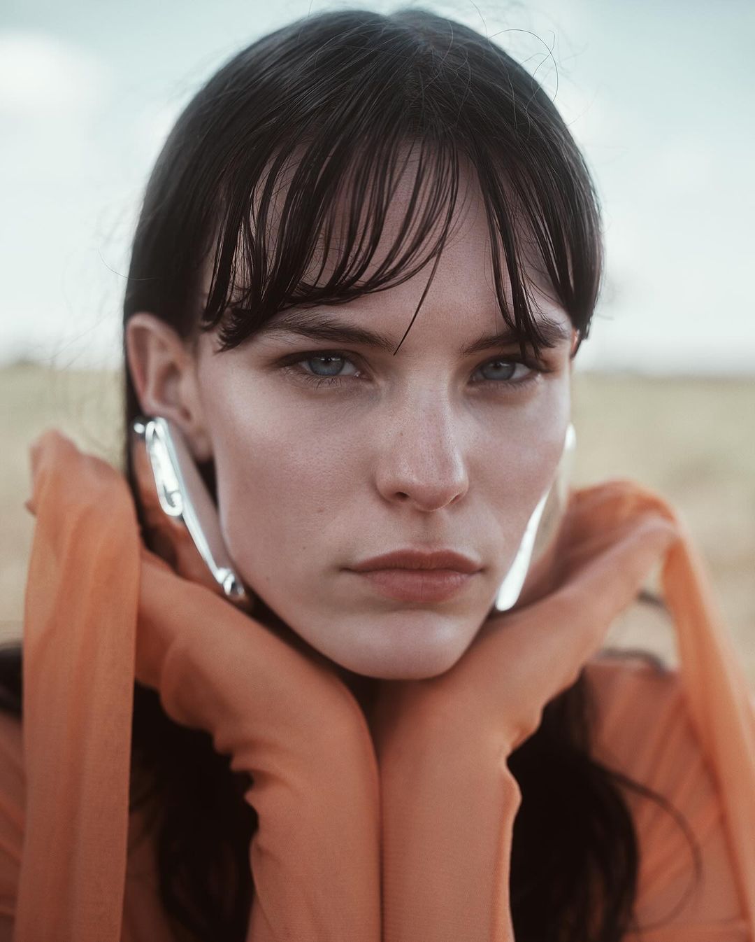 Close-up portrait of a woman with dark hair and bangs wearing orange gloves framing her face, captured by Alessio Albi.