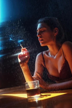 Woman in lingerie holds a lit cigarette in the rain, with a glass of dark liquor on a reflective surface, in a low-light, moody setting.