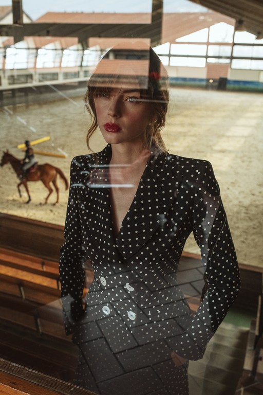Elegant woman in polka dot suit stands indoors with horse and rider visible through a glass partition, creating a layered, stylish composition.