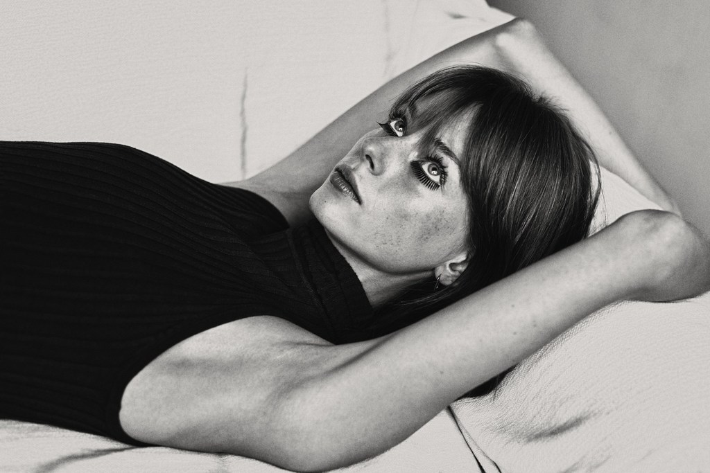 Black and white portrait of a woman lying down with her hands behind her head, looking up with striking eyes and dark hair.