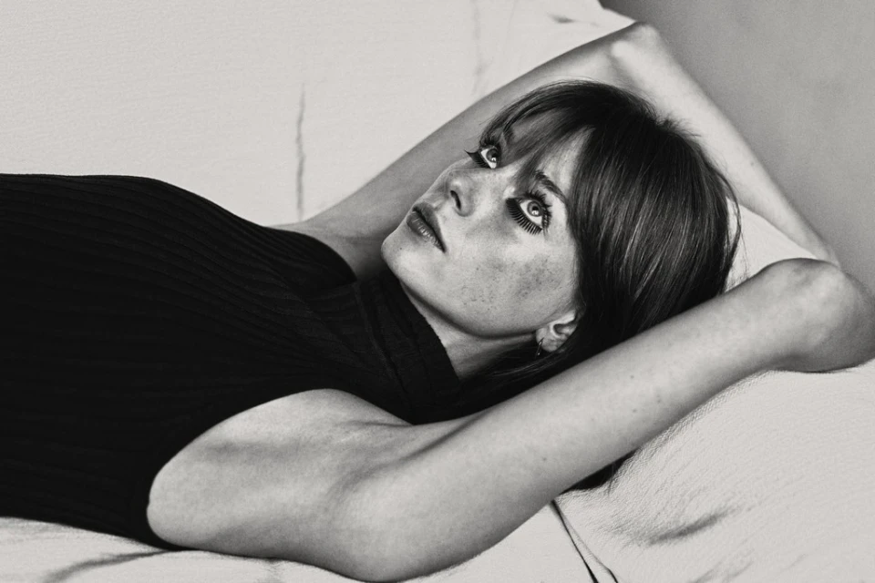 Black and white portrait of a woman lying down with her hands behind her head, looking up with striking eyes and dark hair.