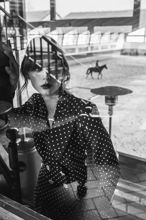 Black and white shot of a woman in a polka dot dress reflected in glass with a horse and rider visible in the background.