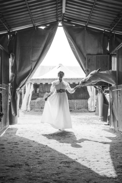 Black and white shot of a woman in a white dress interacting with a white horse inside a stable, illuminated by natural light.