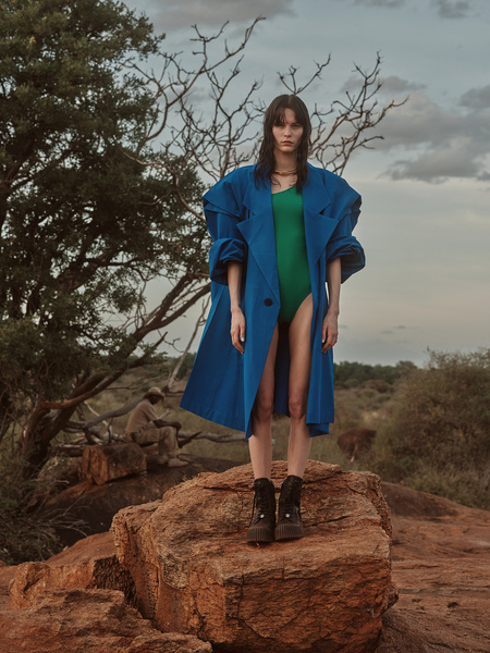 Fashion shot of a young woman posing on a rock formation wearing a bright blue coat and green bodysuit in an outdoor, natural setting.