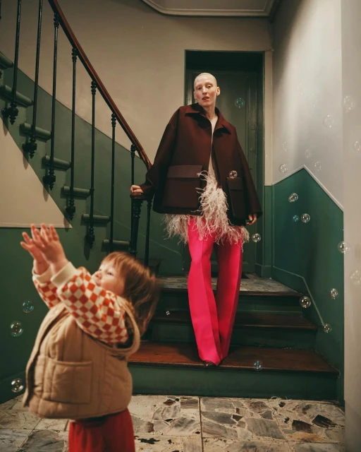 Fashion shot in a stairwell with a child reaching for bubbles and a model standing behind wearing a coat and bright pink pants.