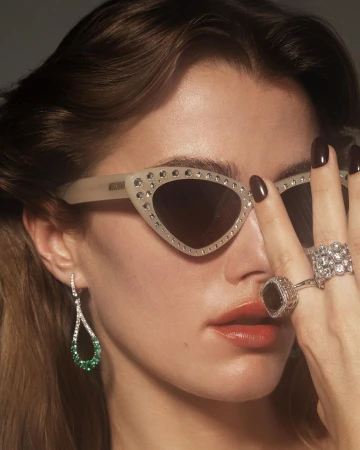 Close-up portrait of a woman wearing jeweled cat-eye sunglasses, sparkling diamond rings, and teardrop emerald earrings.