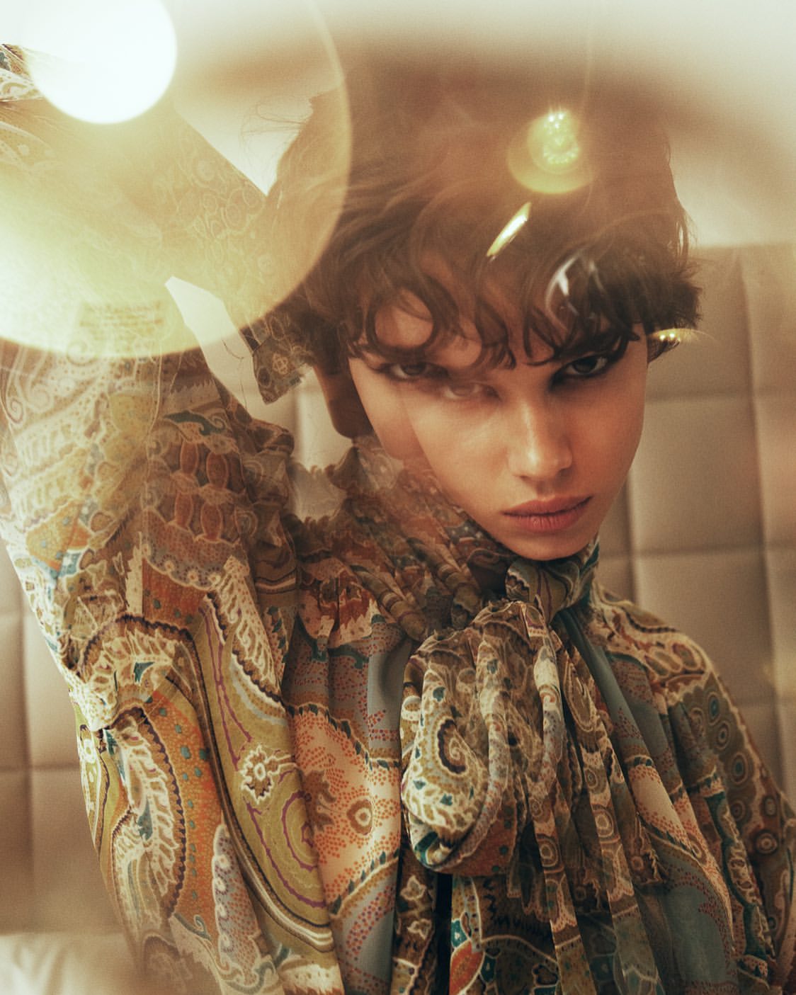 Close-up studio shot of a stylish brunette woman wearing a patterned blouse with a tie, looking at the camera with a neutral expression.