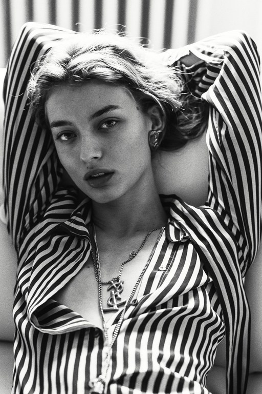Black and white portrait of a young adult woman with blonde hair, wearing a striped shirt and necklace, lying down with arms up.