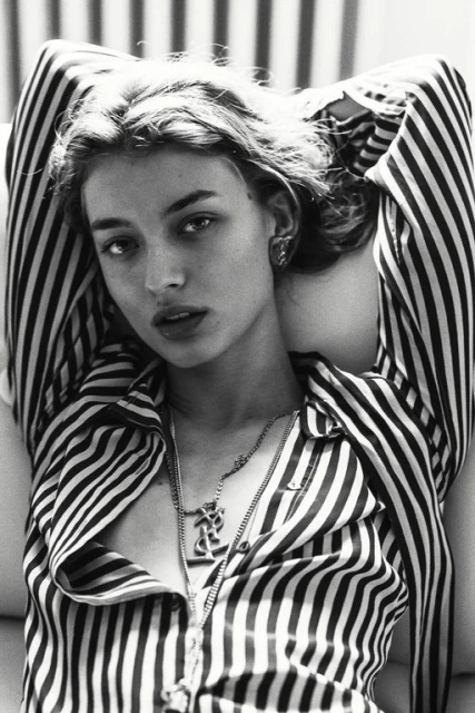Black and white portrait of a young adult woman with blonde hair, wearing a striped shirt and necklace, lying down with arms up.
