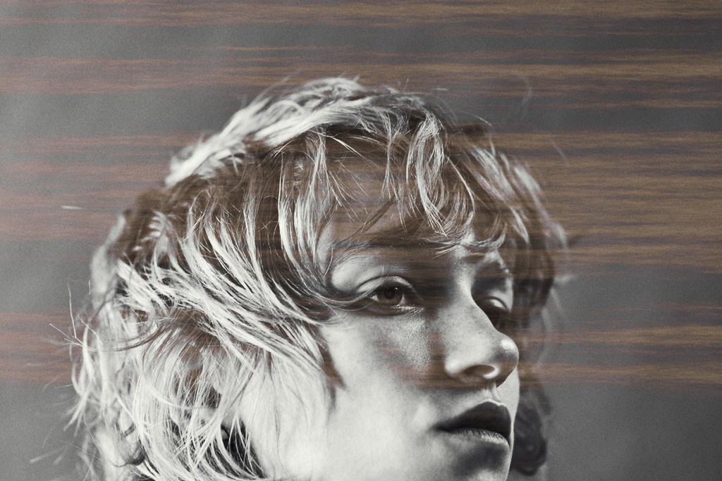 Monochrome portrait of a young adult female with wavy hair and thoughtful expression, overlaid with blurry horizontal wood grain texture.