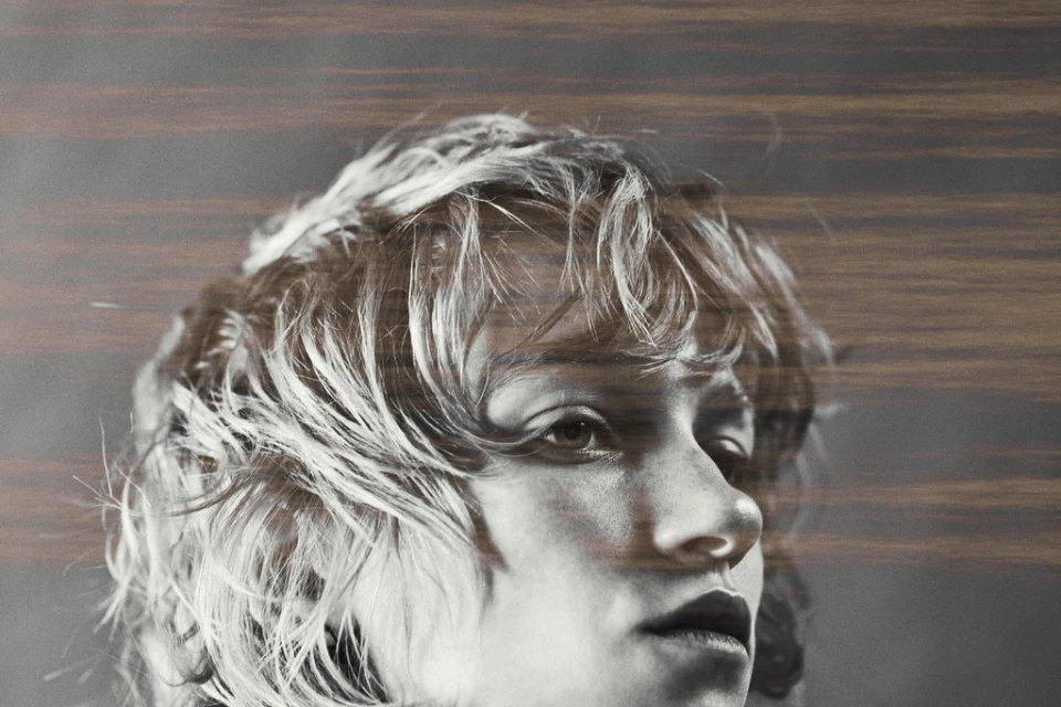 Monochrome portrait of a young adult female with wavy hair and thoughtful expression, overlaid with blurry horizontal wood grain texture.