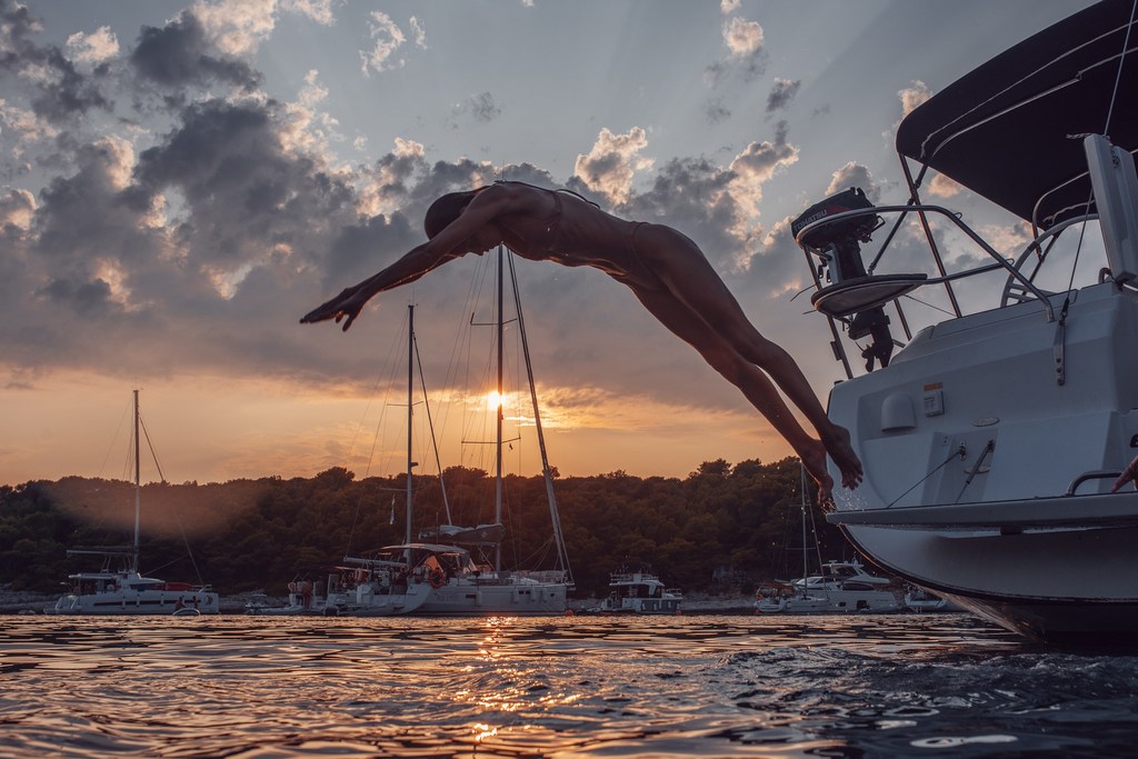 Man dives off white yacht into calm, rippled water during sunset. The yacht is anchored near a green shoreline under a cloudy sky.
