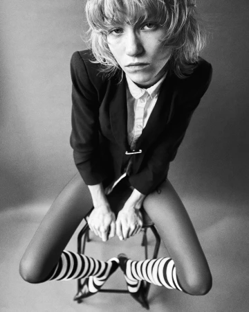 Portrait of a person with short hair wearing a dark blazer over a white shirt and striped socks, seated on a stool.