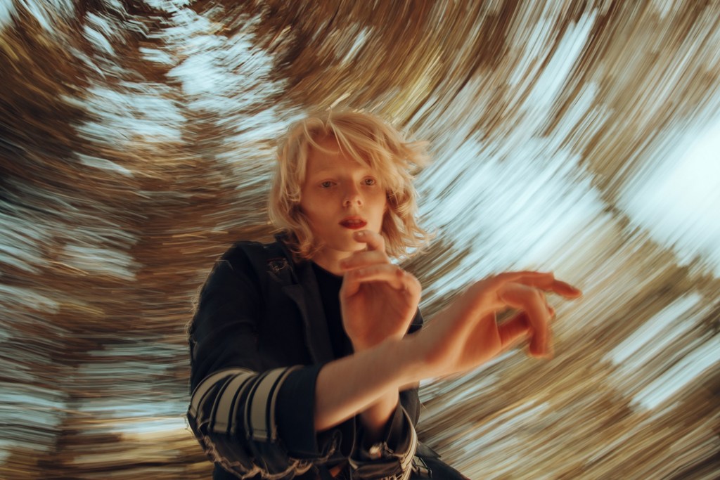 Blonde model in dark jacket with striped sleeve gesturing amid swirling motion blur background by Alessio Albi