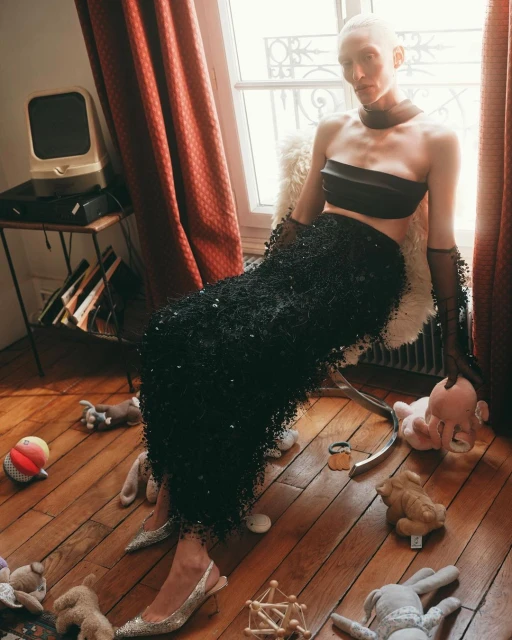 Woman in a black strapless top and beaded skirt poses seated in an indoor setting, surrounded by stuffed animals, near window light.