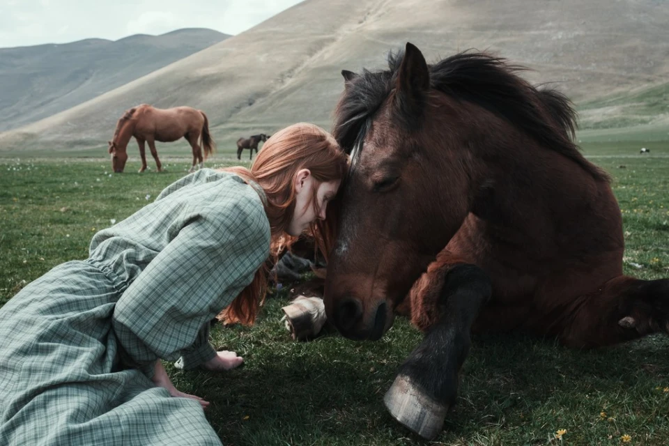 Woman in a green dress kneeling next to a brown horse lying down in a green meadow with mountains in the background in a rural scene.