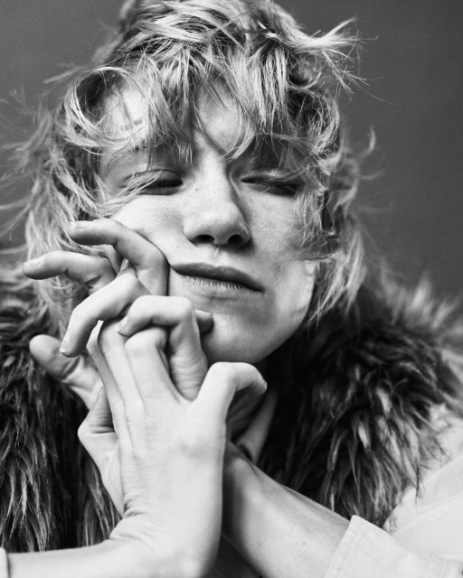 Black and white portrait of an adult with curly blonde hair, hands clasped near the face, wearing a fur collar and white collared shirt.