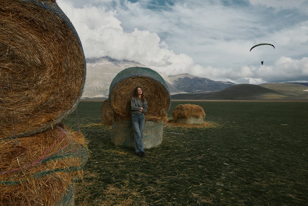 Adult female stands next to round hay bale in open field beneath clouds, with a paraglider visible in the distance.