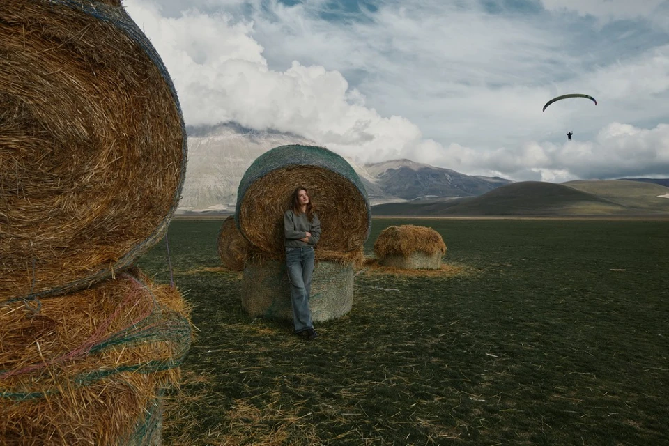 Adult female stands next to round hay bale in open field beneath clouds, with a paraglider visible in the distance.