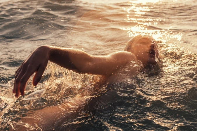 Woman swims in wavy ocean during golden hour. Water splashes around swimmer in horizontal shot emphasizing natural beauty and fitness.