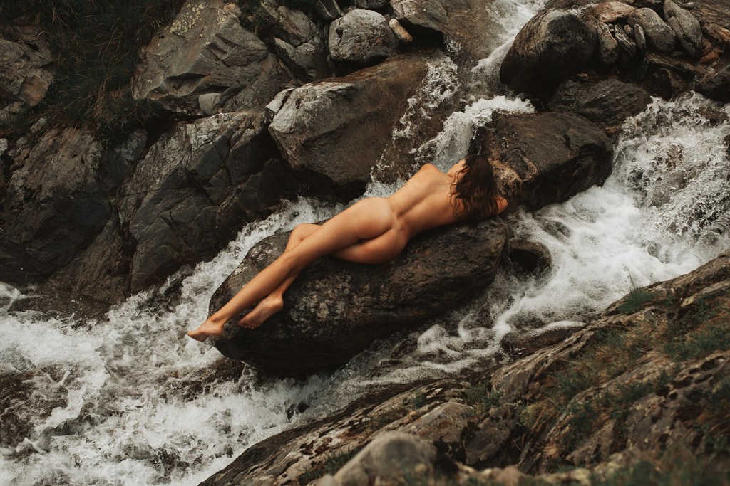 A nude woman lies on a boulder in a river with white water cascading around her and dark rocks and greenery in the background.