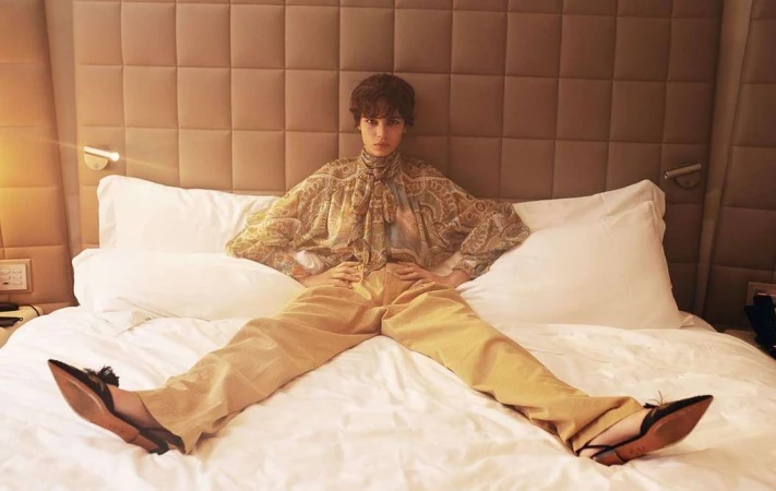 Model with short dark hair seated squarely on a hotel bed wearing a patterned blouse and trousers, photographed by Alessio Albi.