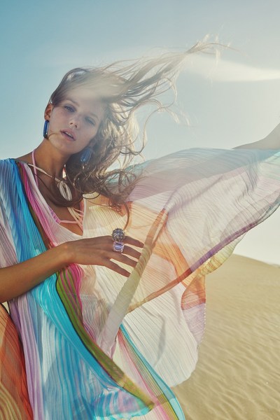 Model in flowing colorful dress posing in sunny desert. She has long hair and wears statement jewelry for a fashion or beauty shoot.
