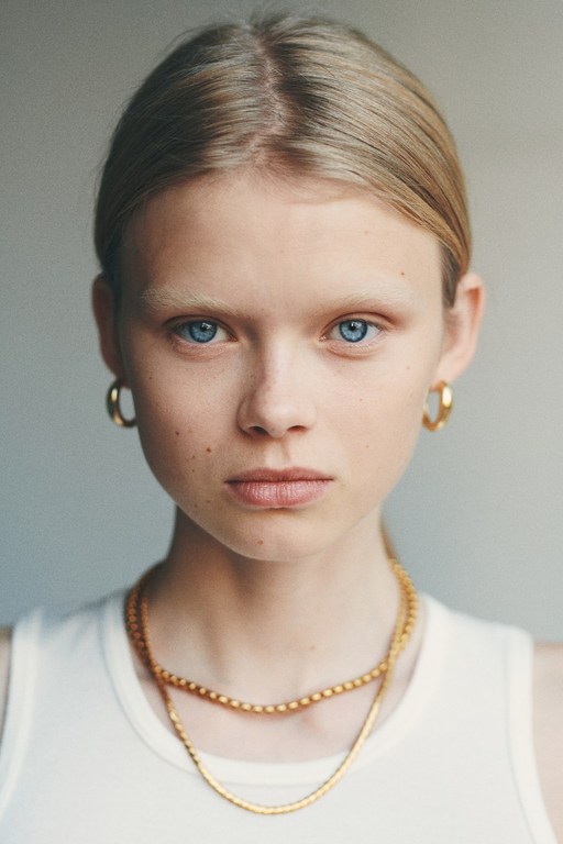 Close-up portrait of a young woman with blonde hair, blue eyes, wearing gold hoop earrings and layered chain necklaces, by Alessio Albi.