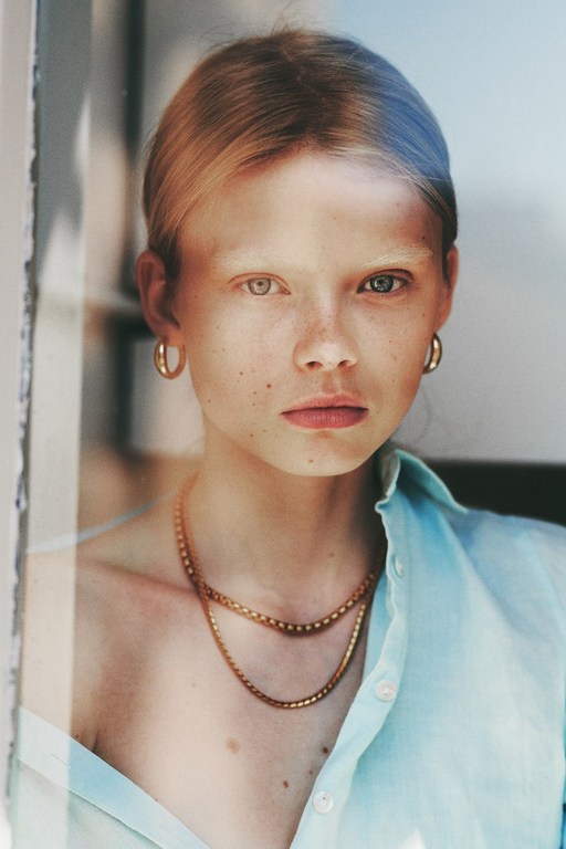 Close-up studio portrait of a young blonde woman with freckles, wearing gold jewelry and a light blue button-down shirt.