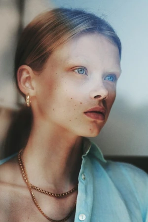 Portrait of a young woman with light hair and blue eyes wearing a light blue buttoned shirt and gold chain necklaces by Alessio Albi.