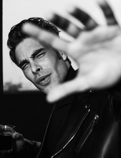 Black and white portrait of a stylish adult man with a hand up towards the camera, wearing a leather jacket and holding a drink.