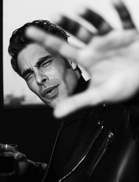 Black and white portrait of a stylish adult man with a hand up towards the camera, wearing a leather jacket and holding a drink.
