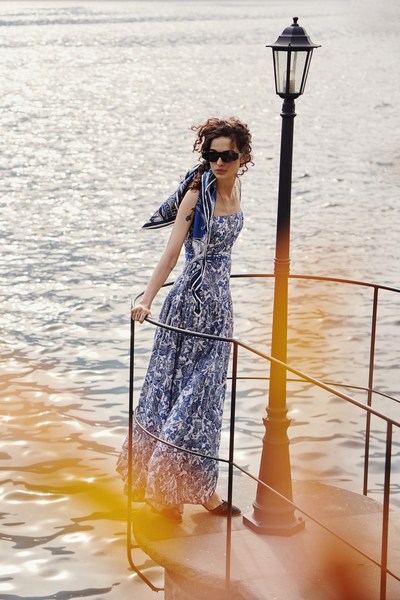 Woman in blue floral dress stands near lamp post by water. She wears sunglasses and a scarf, posing on a sunny day.