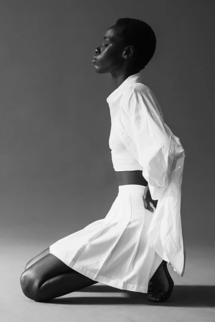 Elegant black woman kneeling in a cropped white shirt and white skirt against a gray backdrop in a studio fashion shot.