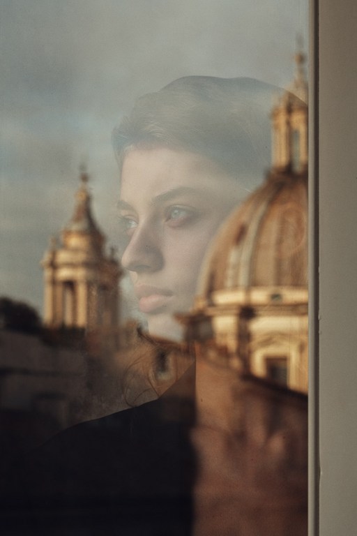 Portrait of a contemplative young woman reflected in a window, with historic architecture visible in the background, bathed in soft light.