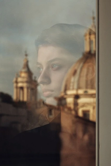 Portrait of a contemplative young woman reflected in a window, with historic architecture visible in the background, bathed in soft light.