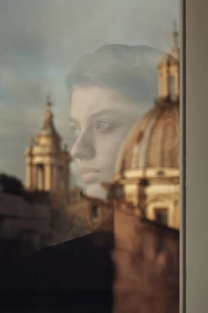 Portrait of a contemplative young woman reflected in a window, with historic architecture visible in the background, bathed in soft light.