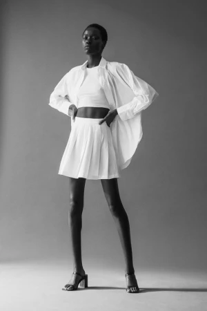 Black and white portrait of a model in a white cropped top, pleated skirt, and oversized shirt by Alessio Albi